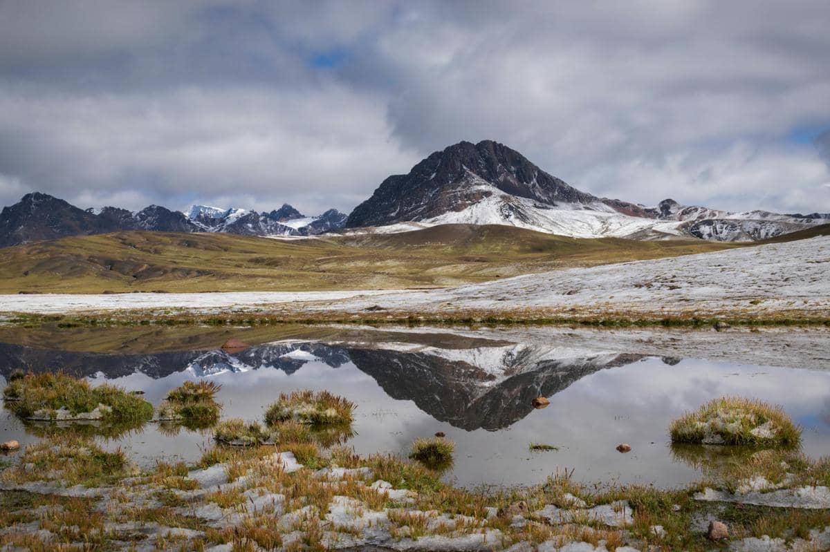 Pemandangan pegunungan Andes dengan puncak bersalju yang terpantul di permukaan danau tenang di bawah langit berawan.