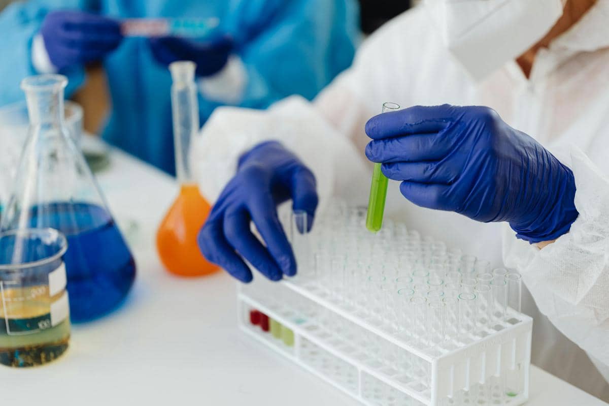 Peneliti mengenakan sarung tangan biru dan pakaian pelindung sedang memegang tabung reaksi berisi cairan hijau di laboratorium.