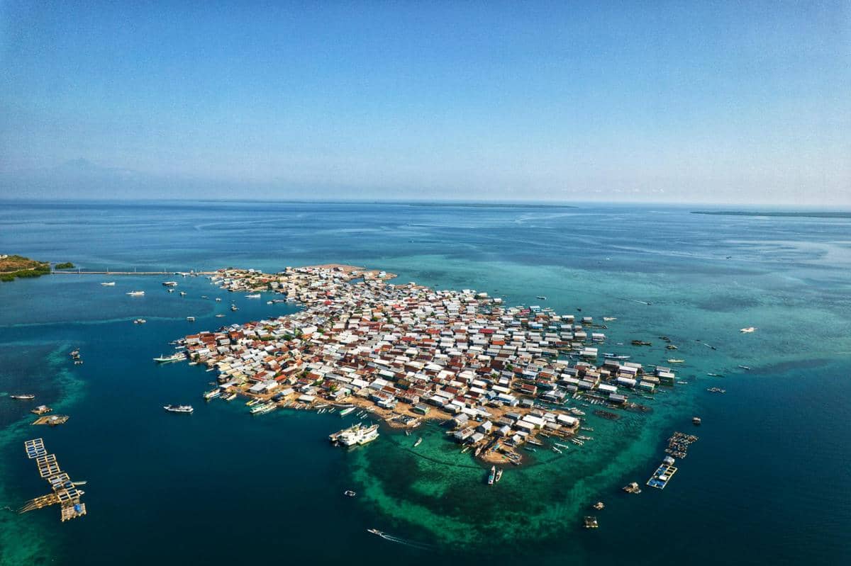 Permukiman padat di tepi laut dengan rumah-rumah berdiri di atas air biru jernih, menggambarkan kehidupan masyarakat pesisir.