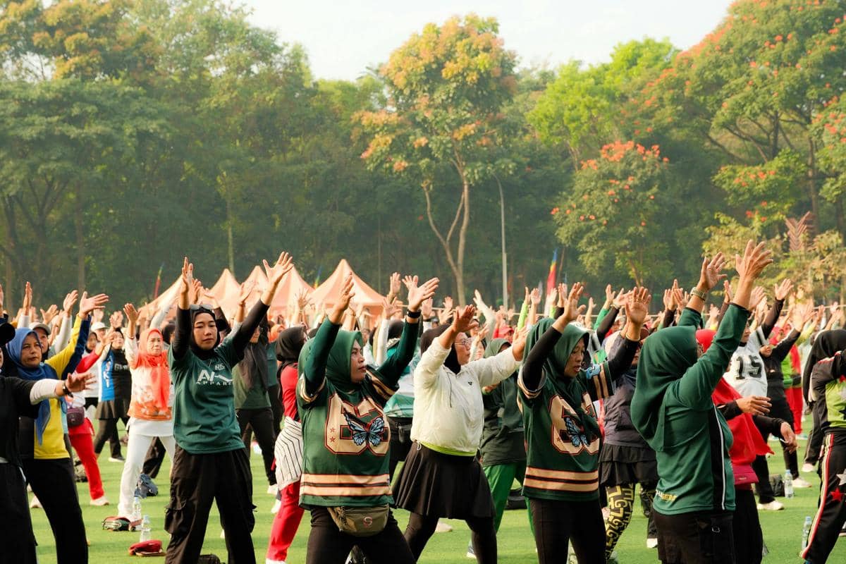 Sekelompok orang melakukan senam aerobik bersama di ruang terbuka hijau dengan latar pepohonan dan suasana pagi yang cerah.