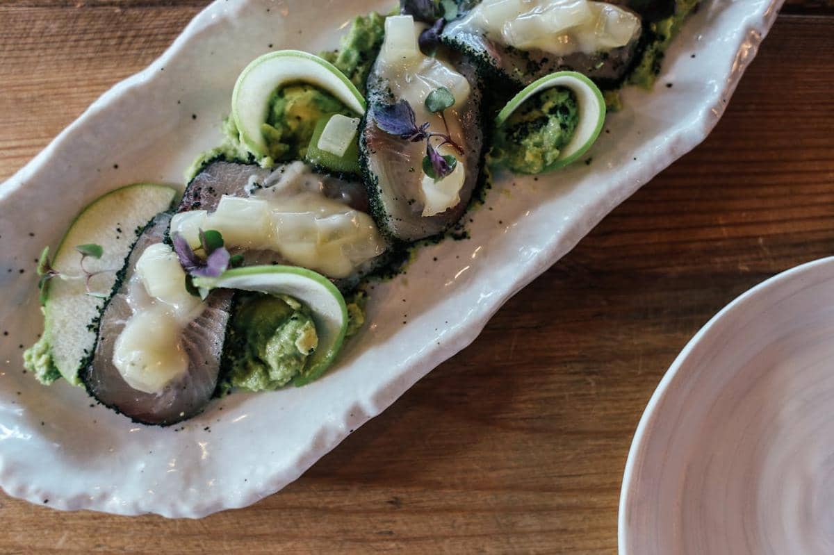 Hidangan poached oyster dan salsify disajikan di atas piring keramik putih dengan hiasan sayuran hijau dan saus lembut.