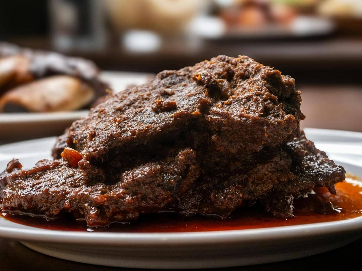 Rendang daging sapi berwarna cokelat pekat dengan kuah kental tersaji di atas piring putih di atas meja makan.