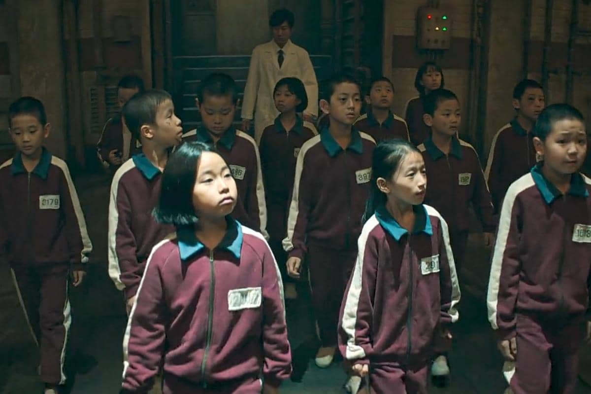 Sekelompok anak mengenakan seragam olahraga merah marun dengan nomor identitas berjalan di koridor gelap bersama seorang pria berjas putih di belakang mereka.