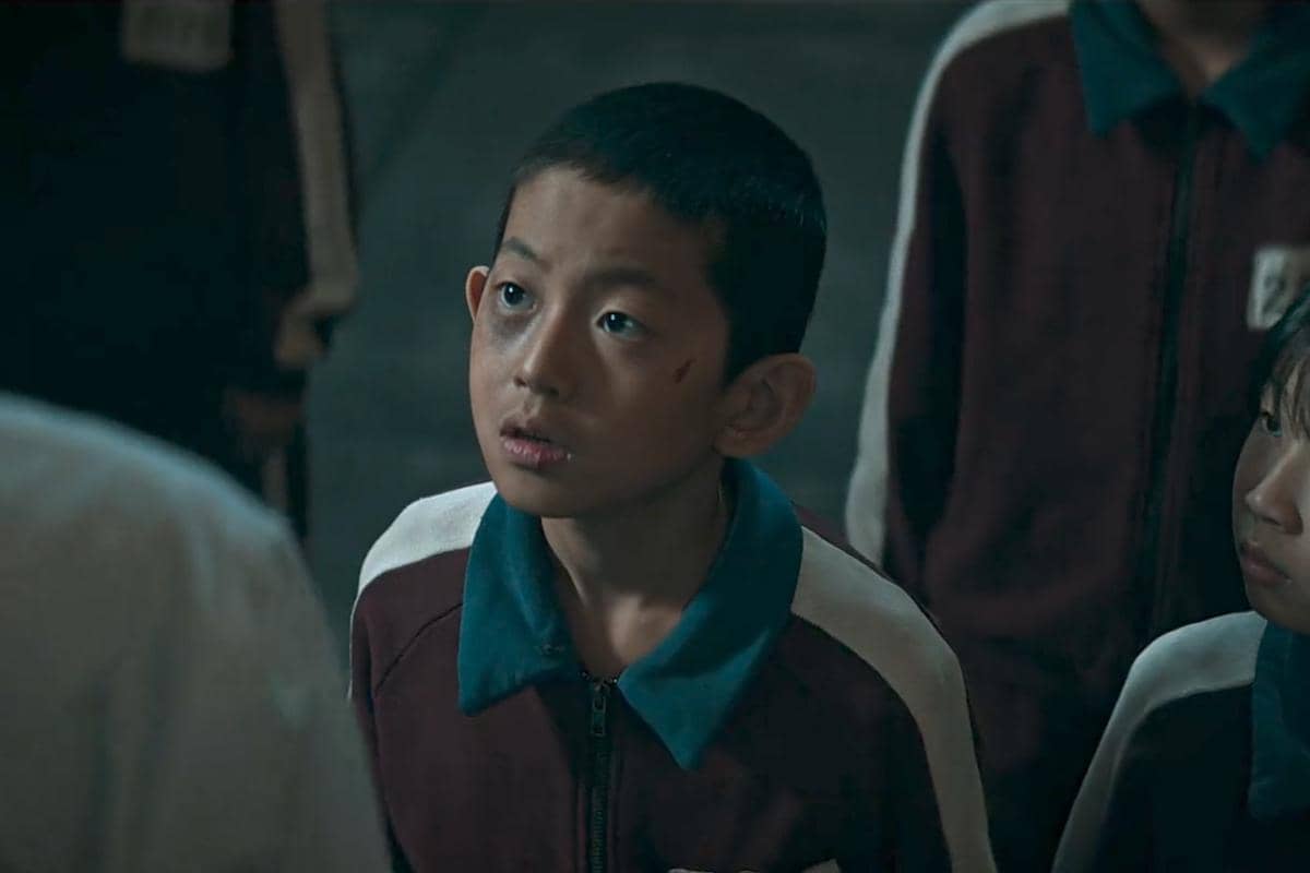 Seorang anak laki-laki mengenakan jaket olahraga merah marun dengan kerah biru, menatap ke atas dengan ekspresi serius dalam adegan drama The Wonderfools.