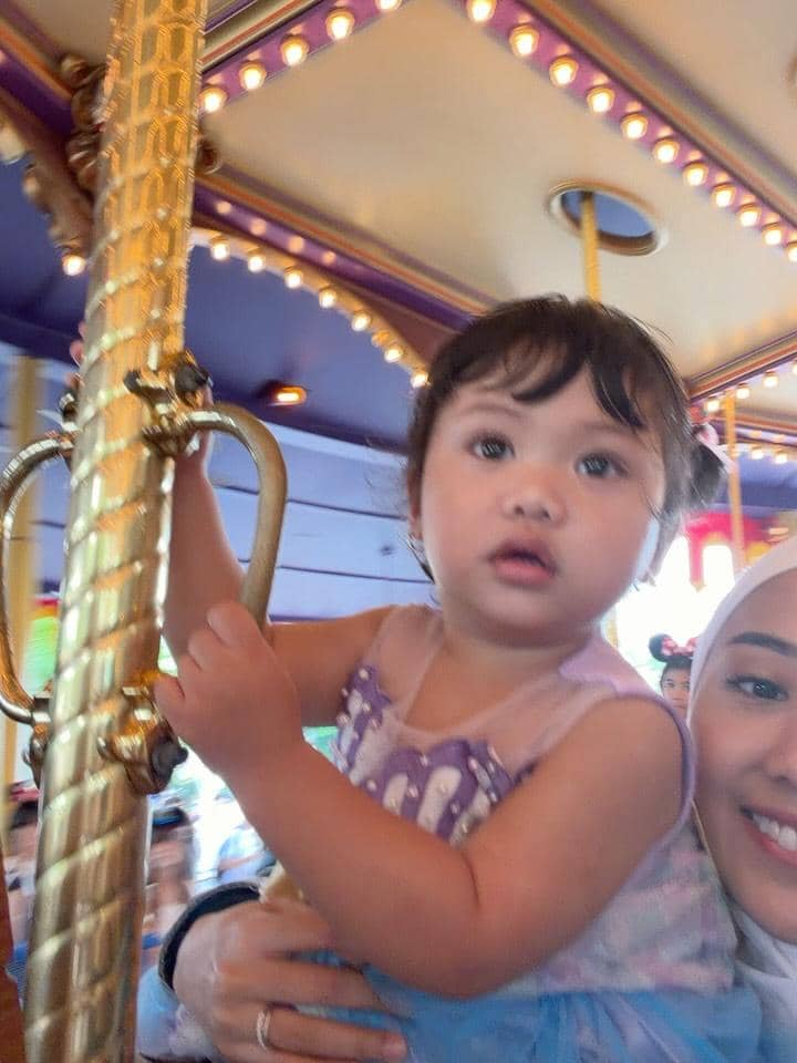 Seorang anak kecil mengenakan gaun ungu sedang bermain di wahana komidi putar bersama seorang wanita di Disneyland Hong Kong.