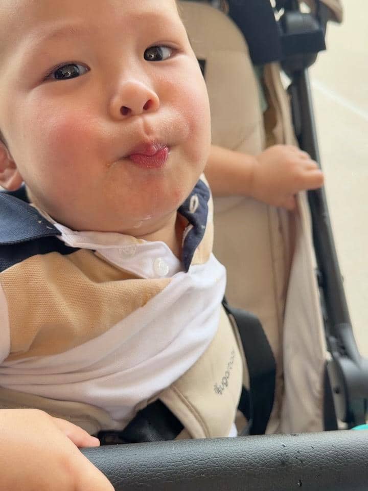 Seorang balita duduk di kereta dorong dengan ekspresi lucu sambil mengerucutkan bibir, mengenakan kaus berkerah warna krem dan putih.