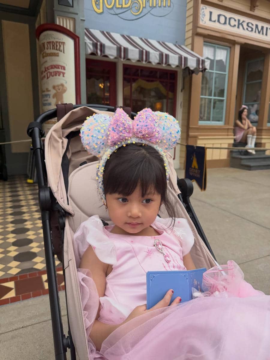 Seorang anak kecil mengenakan gaun berwarna merah muda dan bando telinga Minnie Mouse duduk di kereta dorong di area Disneyland Hong Kong.