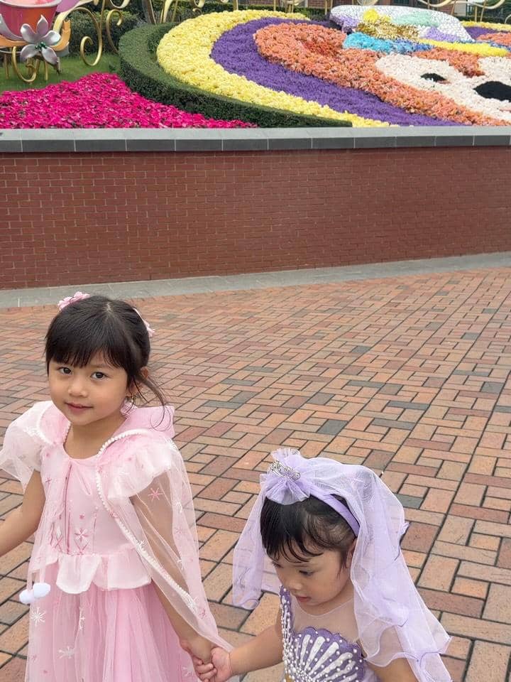 Dua anak kecil mengenakan kostum putri berwarna pink dan ungu berjalan bergandengan tangan di area Disneyland Hong Kong.