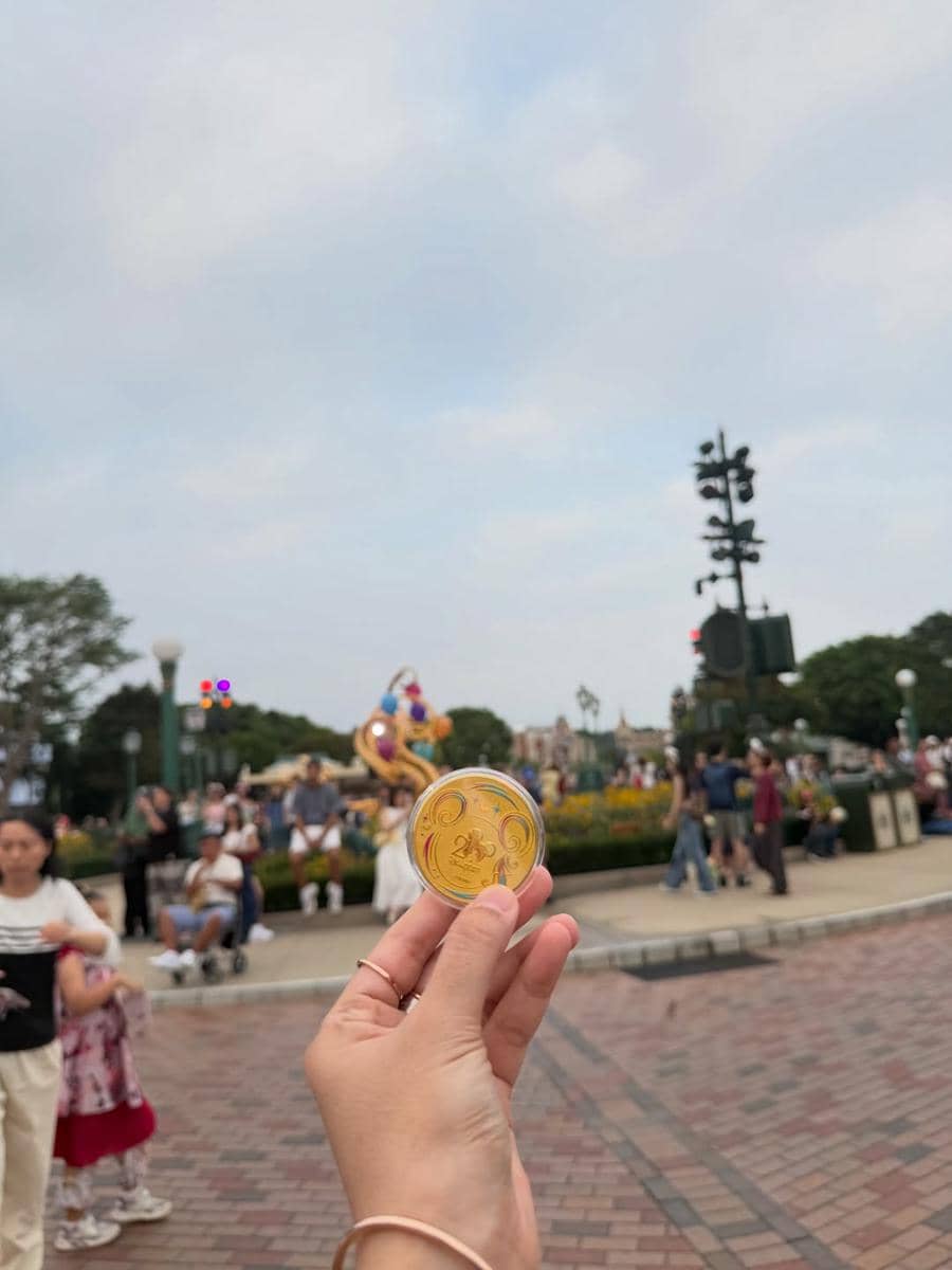 Tangan memegang koin suvenir berwarna emas dengan latar suasana ramai pengunjung di area Disneyland Hong Kong.