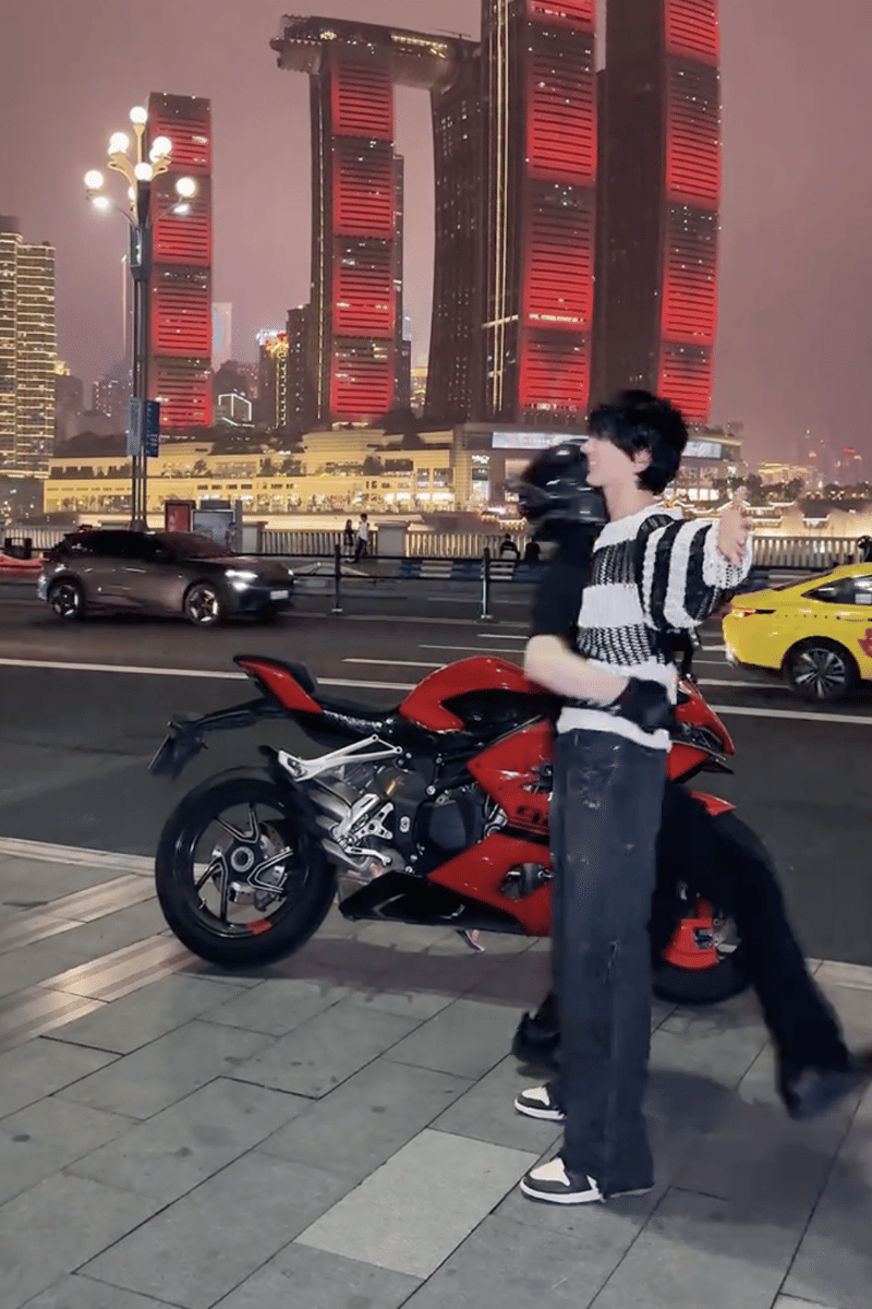 Seorang pria mengenakan sweter hitam putih berpose di depan motor sport merah di jalan kota dengan gedung tinggi bercahaya merah di latar belakang.