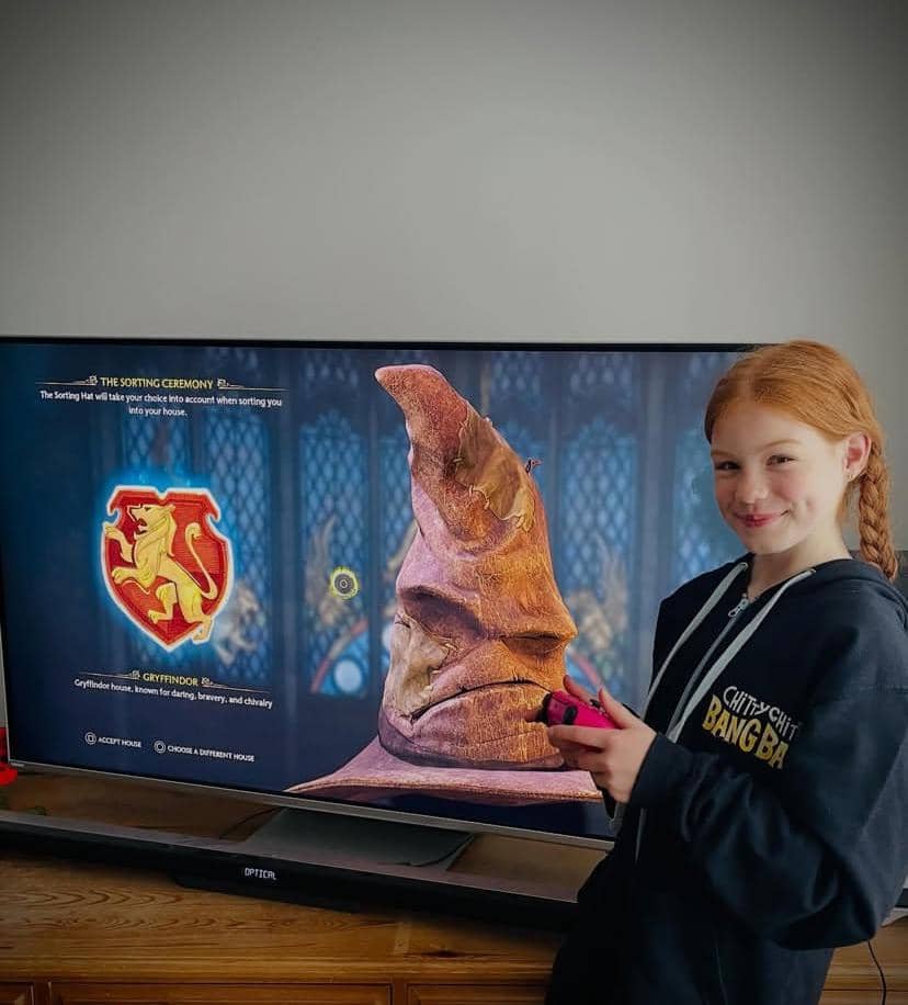 Seorang anak perempuan tersenyum sambil memegang kontroler gim di depan layar televisi yang menampilkan topi penyortiran dari dunia Harry Potter.