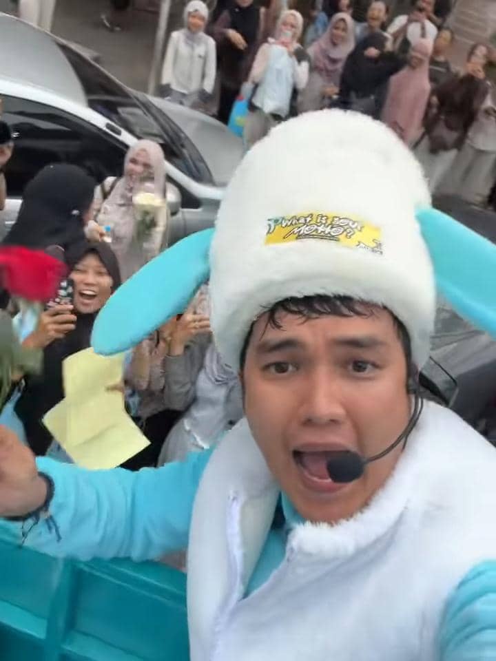 Seorang pria mengenakan kostum topi berbentuk telinga biru dan mikrofon headset berpose di depan kerumunan penggemar yang antusias di jalan.