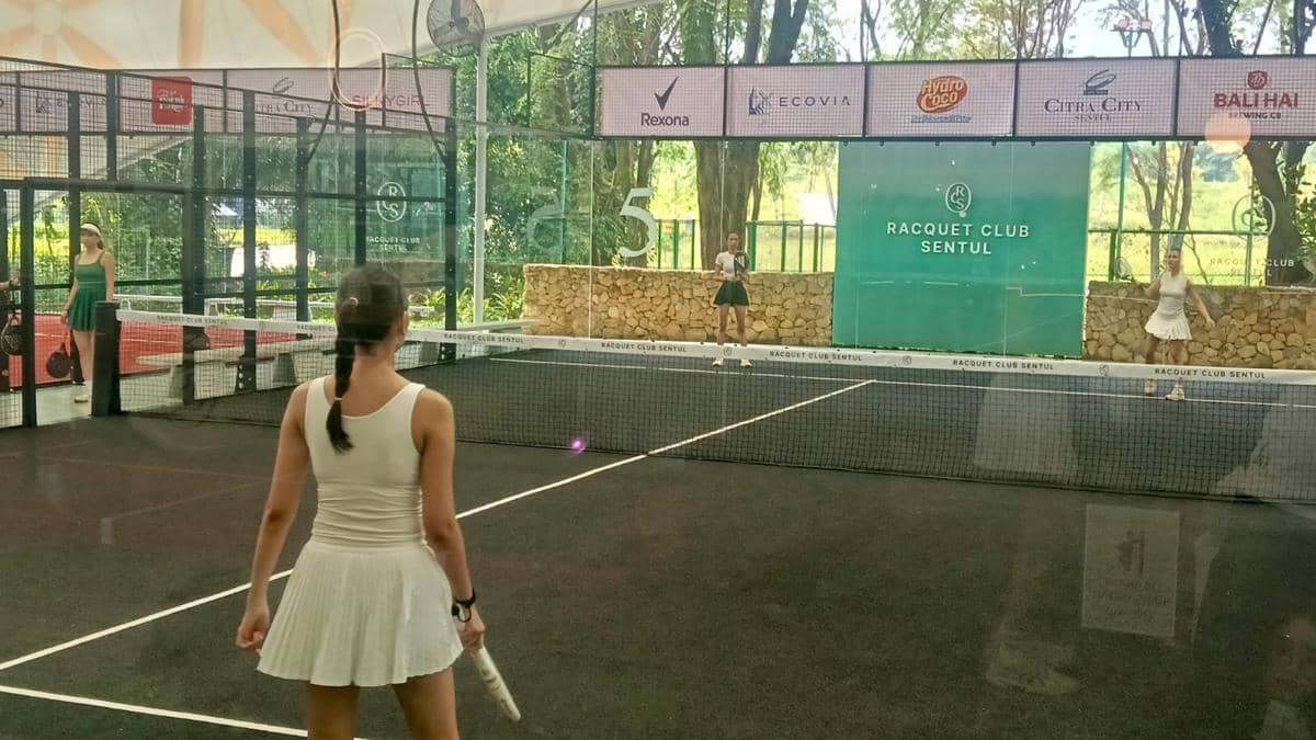 Beberapa pemain sedang bertanding di lapangan padel modern Racquet Club Sentul yang dikelilingi dinding kaca dan pepohonan hijau.