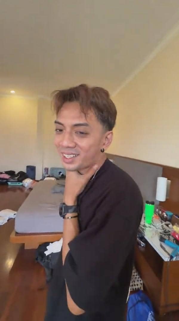 Seorang pria tersenyum sambil memegang lehernya di kamar dengan tempat tidur dan meja yang berantakan di latar belakang.