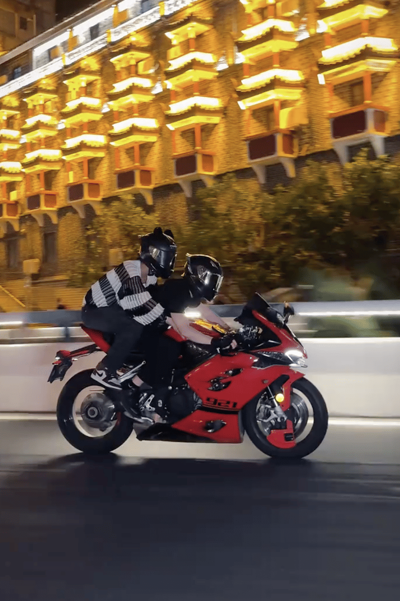 Dua orang mengenakan helm hitam mengendarai motor sport merah di jalan malam dengan latar gedung bercahaya di China.