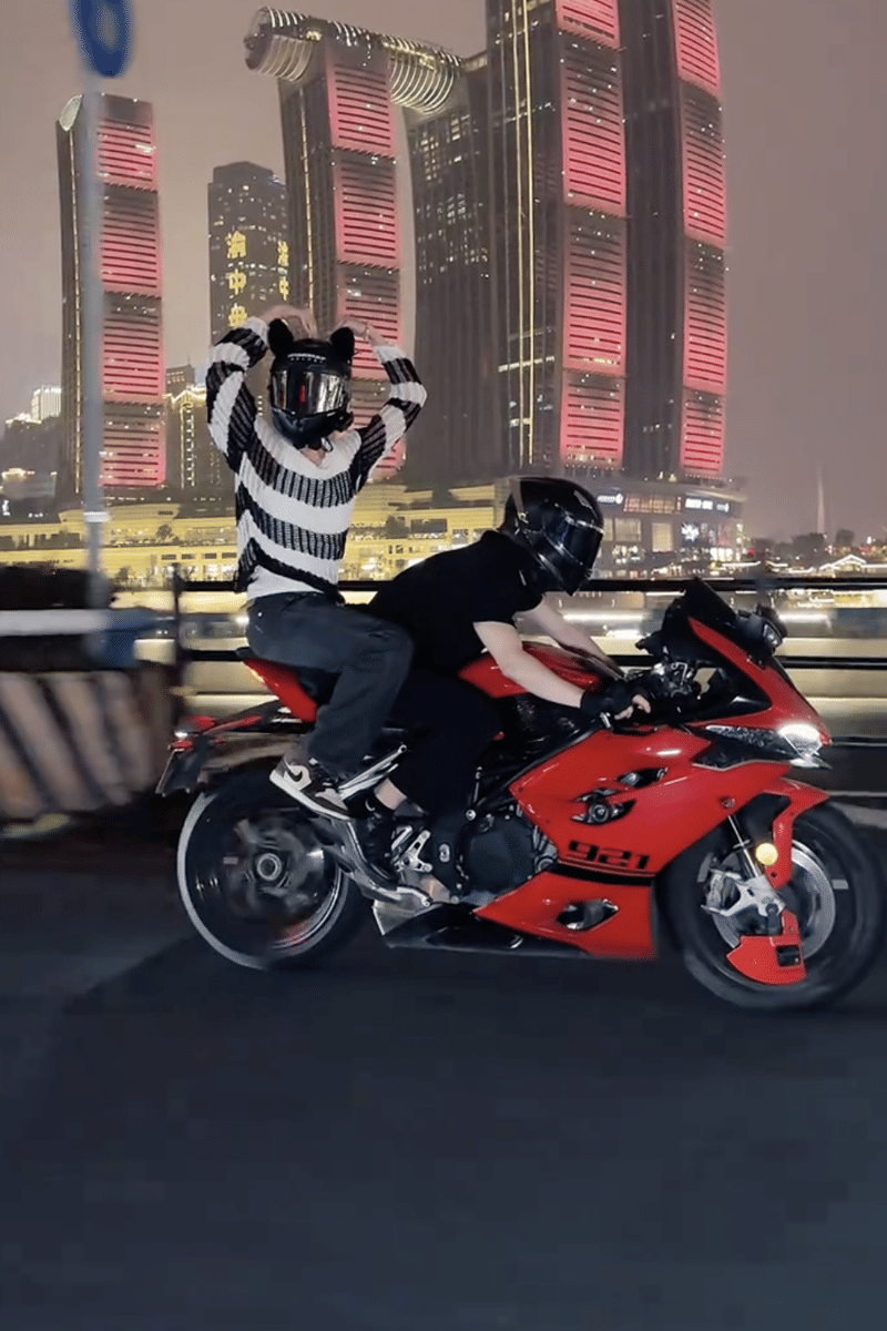 Dua orang mengenakan helm hitam mengendarai motor sport merah di malam hari dengan latar gedung tinggi bercahaya merah di China.