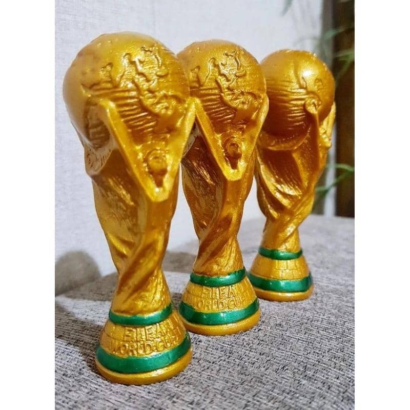 Tiga miniatur trofi Piala Dunia berwarna emas dengan dasar hijau tersusun rapi di atas permukaan kain abu-abu.