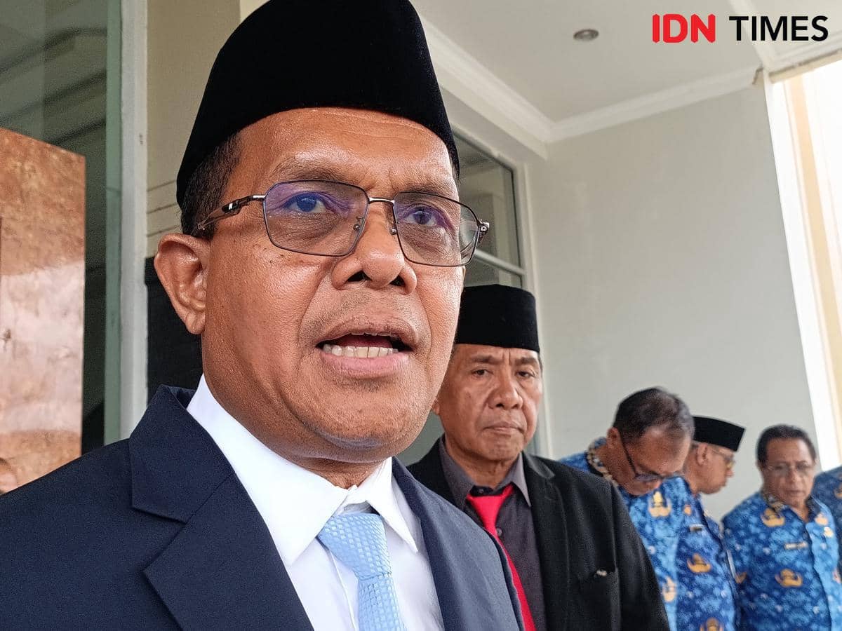 Seorang pria mengenakan jas hitam, dasi biru muda, dan peci hitam berbicara di depan gedung dengan beberapa orang berdiri di belakangnya.