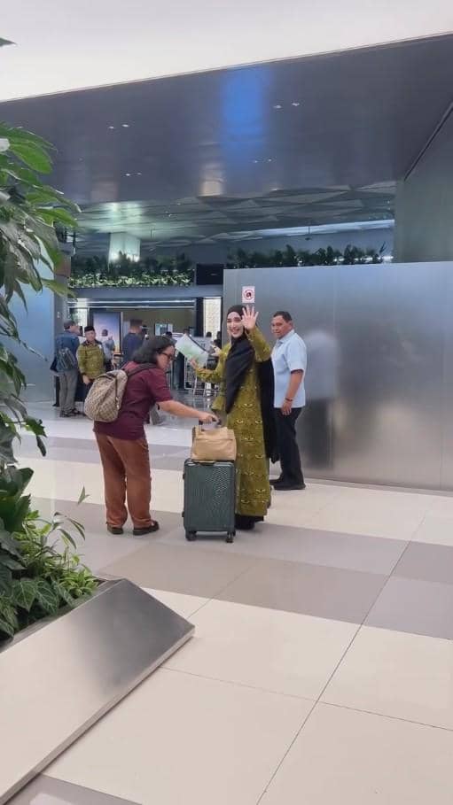 Seorang wanita mengenakan pakaian hijau dan kerudung hitam melambaikan tangan di bandara sambil membawa koper bersama seorang pria.