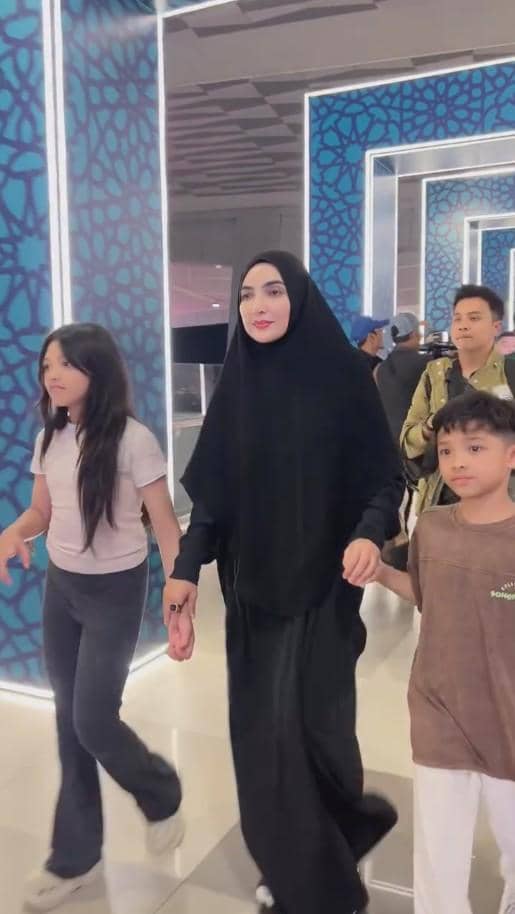Seorang perempuan berhijab hitam berjalan di bandara sambil menggandeng dua anak, dengan latar dinding bermotif biru bercahaya.