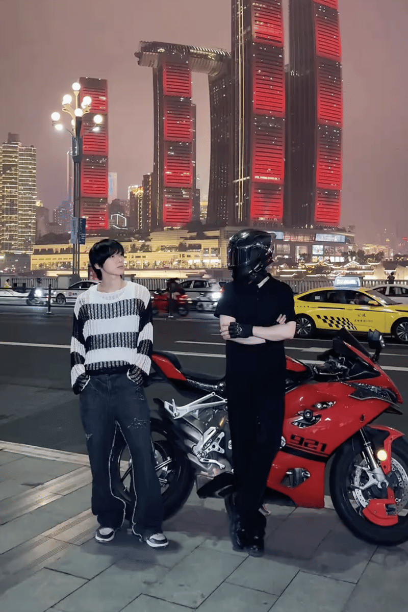 Dua orang berpose di tepi jalan malam hari dengan latar gedung tinggi bercahaya merah dan motor sport merah di China.