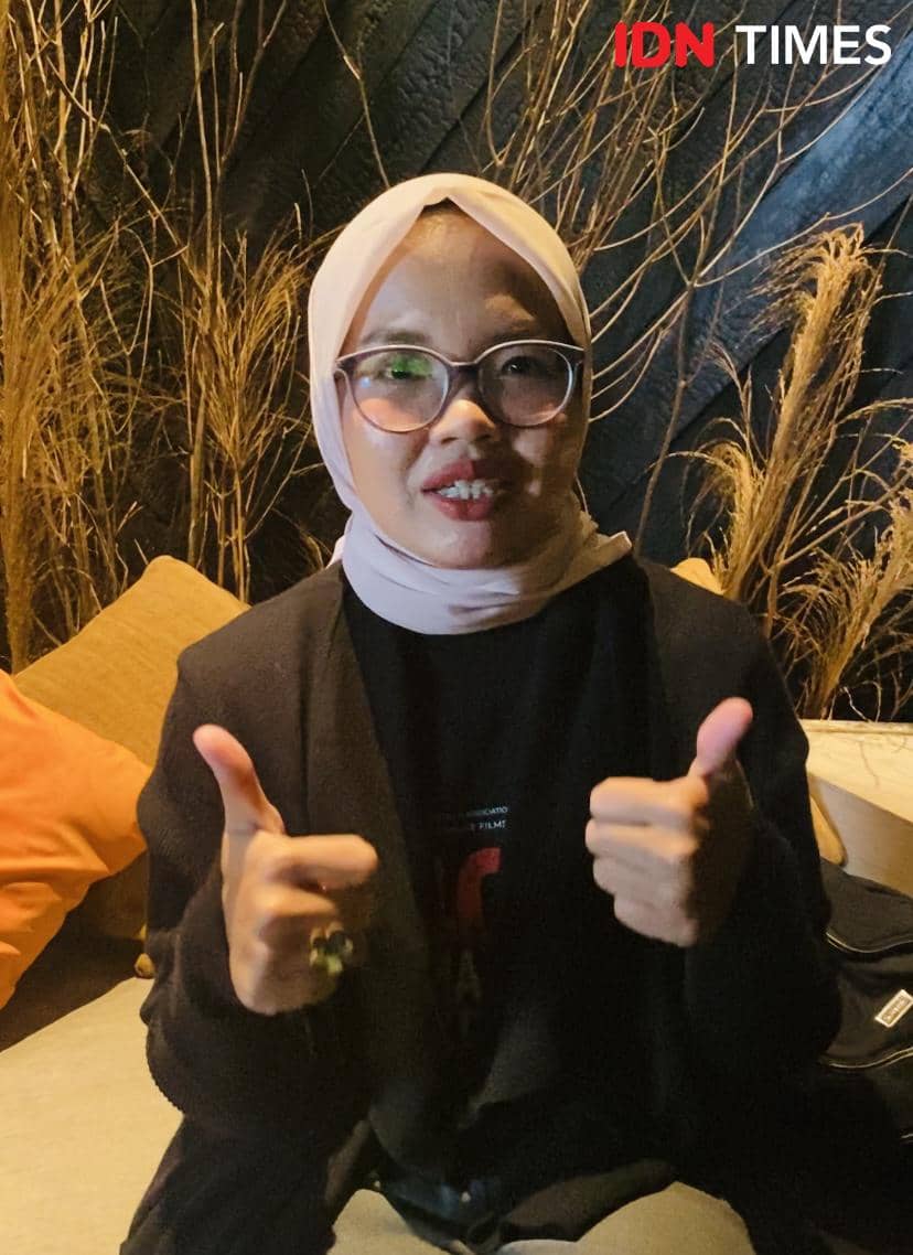 Seorang perempuan berhijab dan berkacamata duduk di sofa dengan latar dekorasi ranting kering sambil mengacungkan dua jempol.