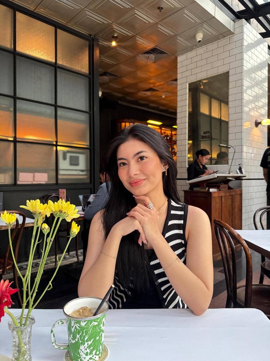 Seorang perempuan berambut panjang duduk di meja kafe dengan minuman dan bunga di depannya, mengenakan atasan hitam bergaris putih.