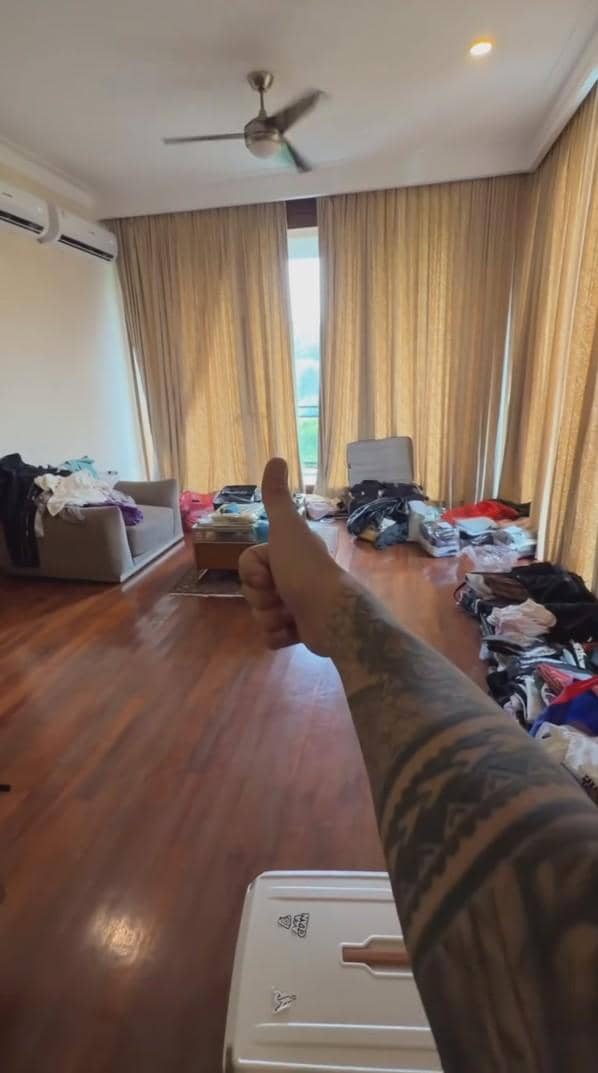Sebuah ruangan dengan lantai kayu, tirai tertutup, koper terbuka, dan pakaian berserakan, dengan tangan bertato mengacungkan jempol di depan kamera.