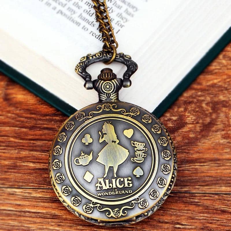 Jam saku antik berwarna perunggu dengan ukiran karakter dan tulisan Alice in Wonderland, diletakkan di atas meja kayu dekat buku terbuka.