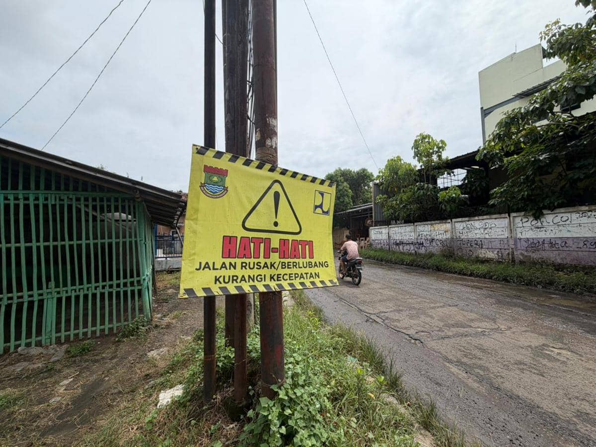 Papan peringatan bertuliskan hati-hati jalan rusak dan berlubang terpasang di tepi Jalan Rajeg-Mauk dengan pengendara motor melintas.
