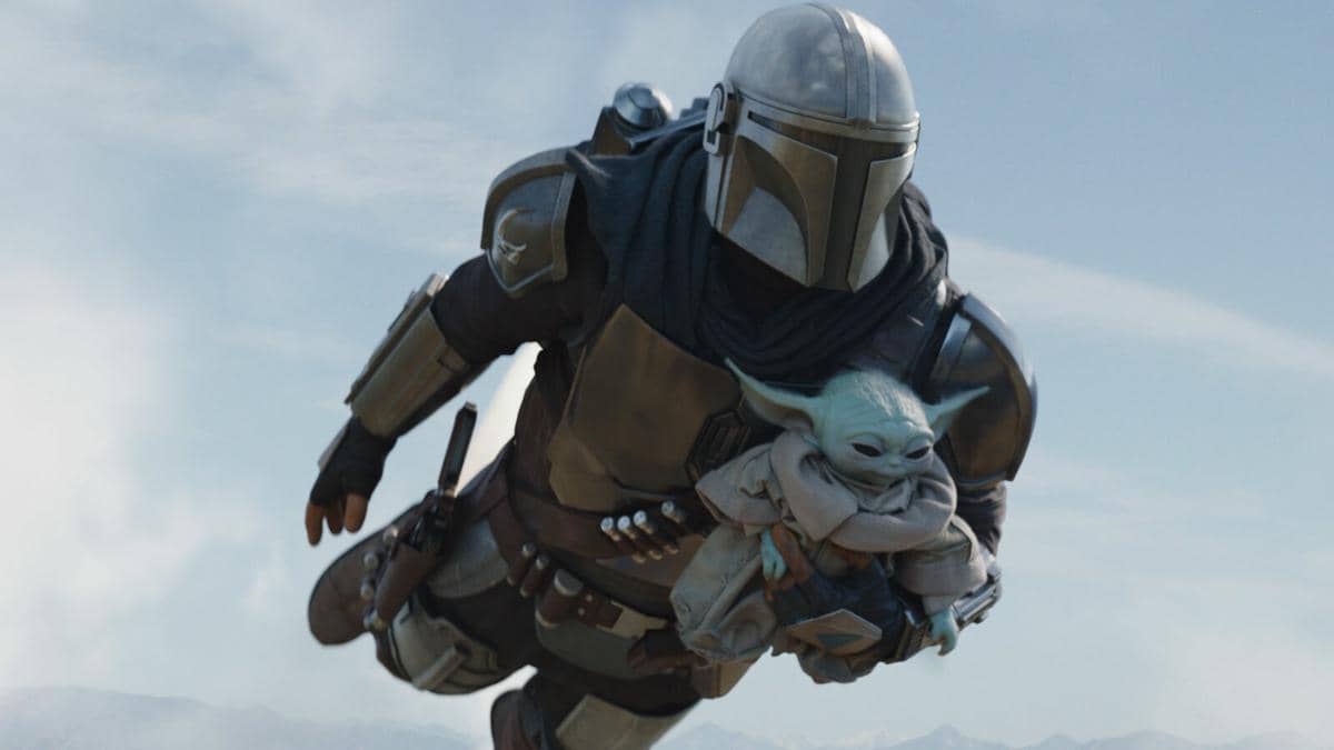 Karakter Din Djarin mengenakan baju besi Mandalorian terbang di udara sambil membawa Grogu di lengannya dengan latar langit biru.