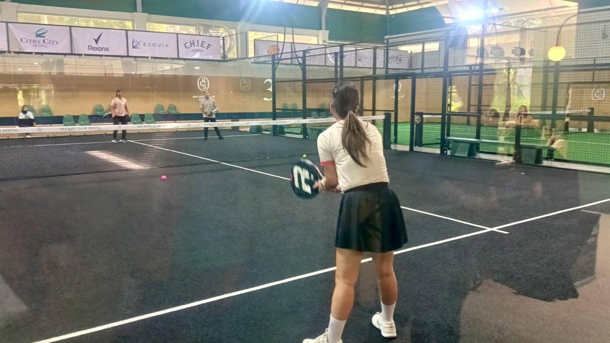 Seorang wanita bersiap memukul bola di lapangan padel indoor Racquet Club Sentul dengan beberapa pemain lain di seberang net.