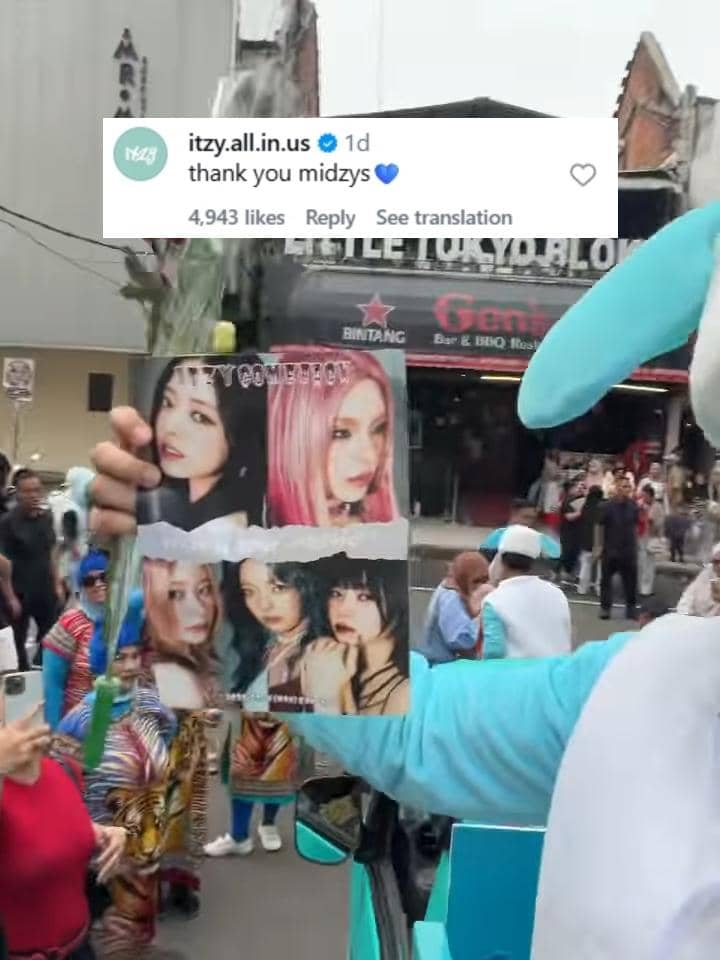 Seseorang mengenakan kostum biru memegang poster grup musik wanita Korea di area ramai dengan komentar akun resmi ITZY di layar.