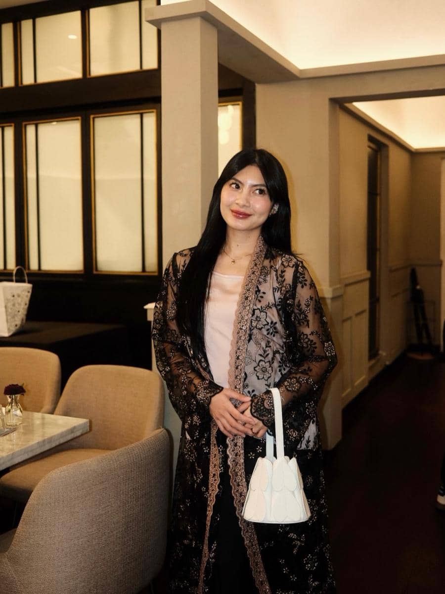 Seorang wanita berambut panjang mengenakan kebaya hitam bermotif renda dan membawa tas putih kecil di dalam ruangan bergaya modern.