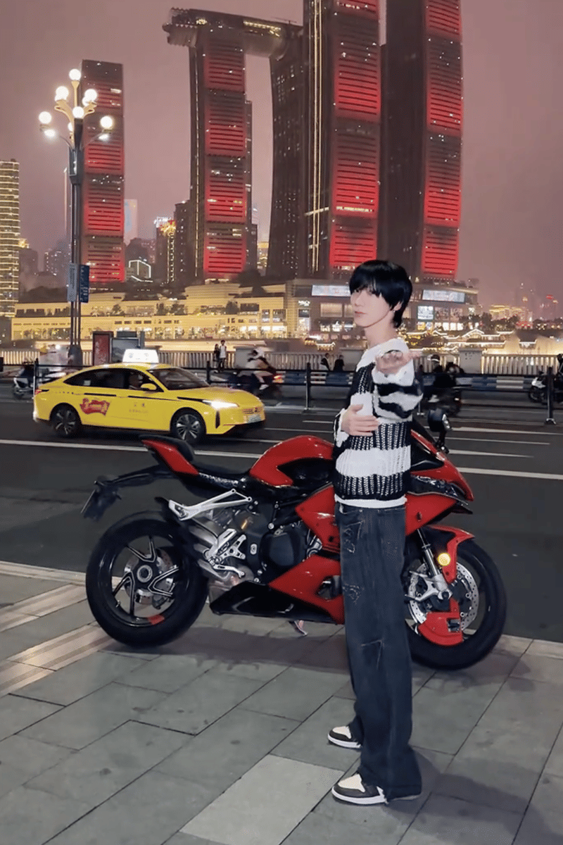 Seorang pria berpose di depan motor sport merah di jalan kota dengan gedung tinggi bercahaya merah di latar belakang malam hari.