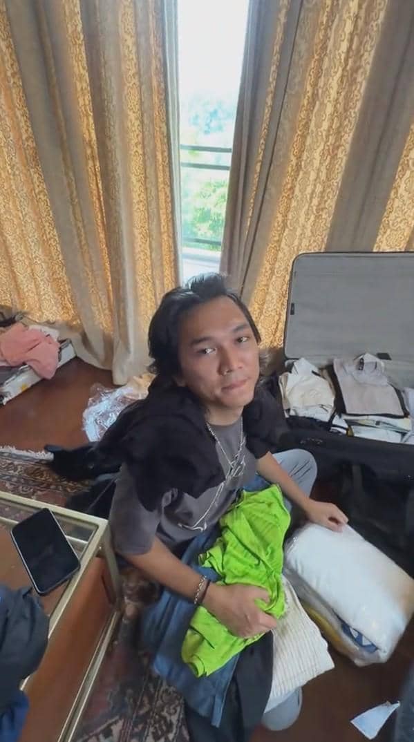 Seorang pria duduk di lantai kamar dengan koper terbuka di sampingnya, memegang pakaian berwarna hijau di tangan.