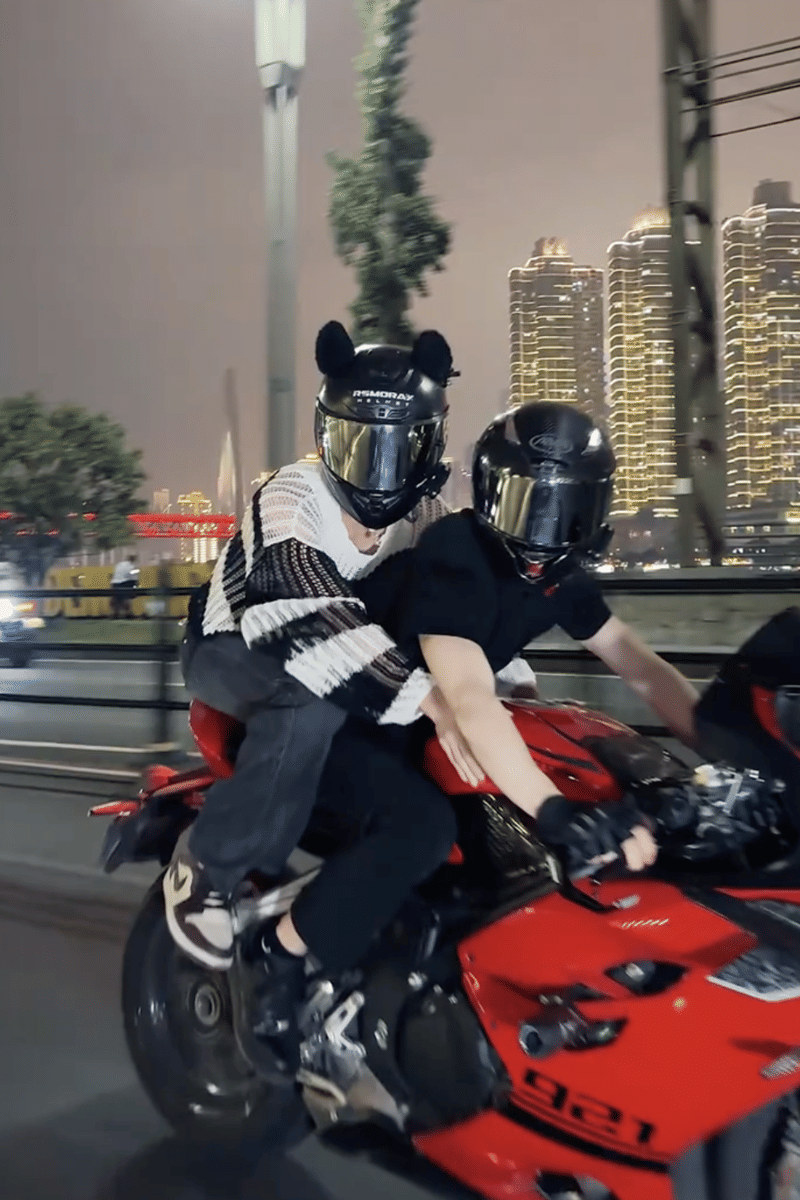 Dua orang mengenakan helm hitam sedang mengendarai motor sport merah di jalan kota pada malam hari dengan gedung tinggi bercahaya di latar belakang.