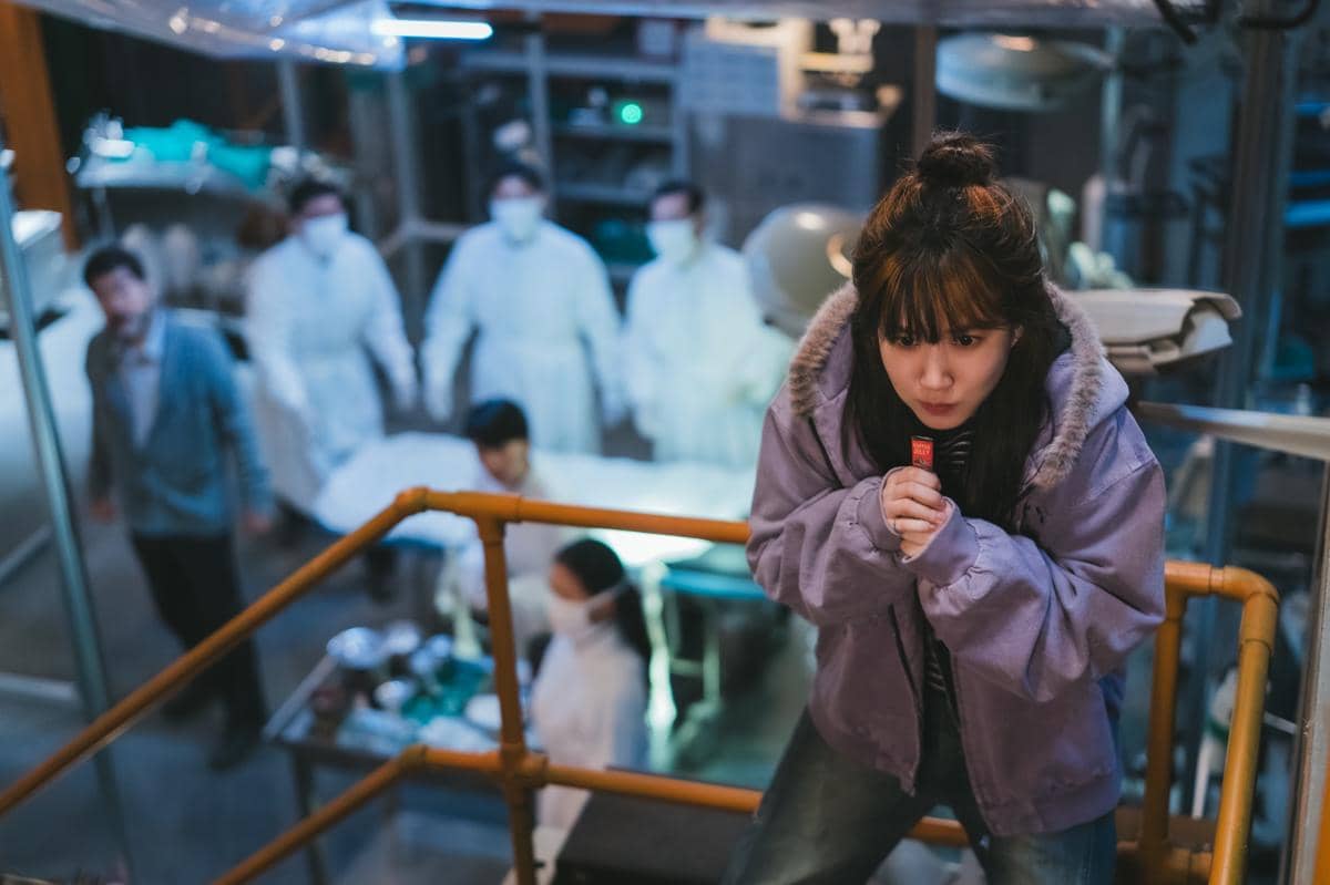Seorang perempuan berjaket ungu berdiri di atas tangga logam dengan ekspresi tegang, sementara beberapa orang berseragam laboratorium berada di belakangnya.