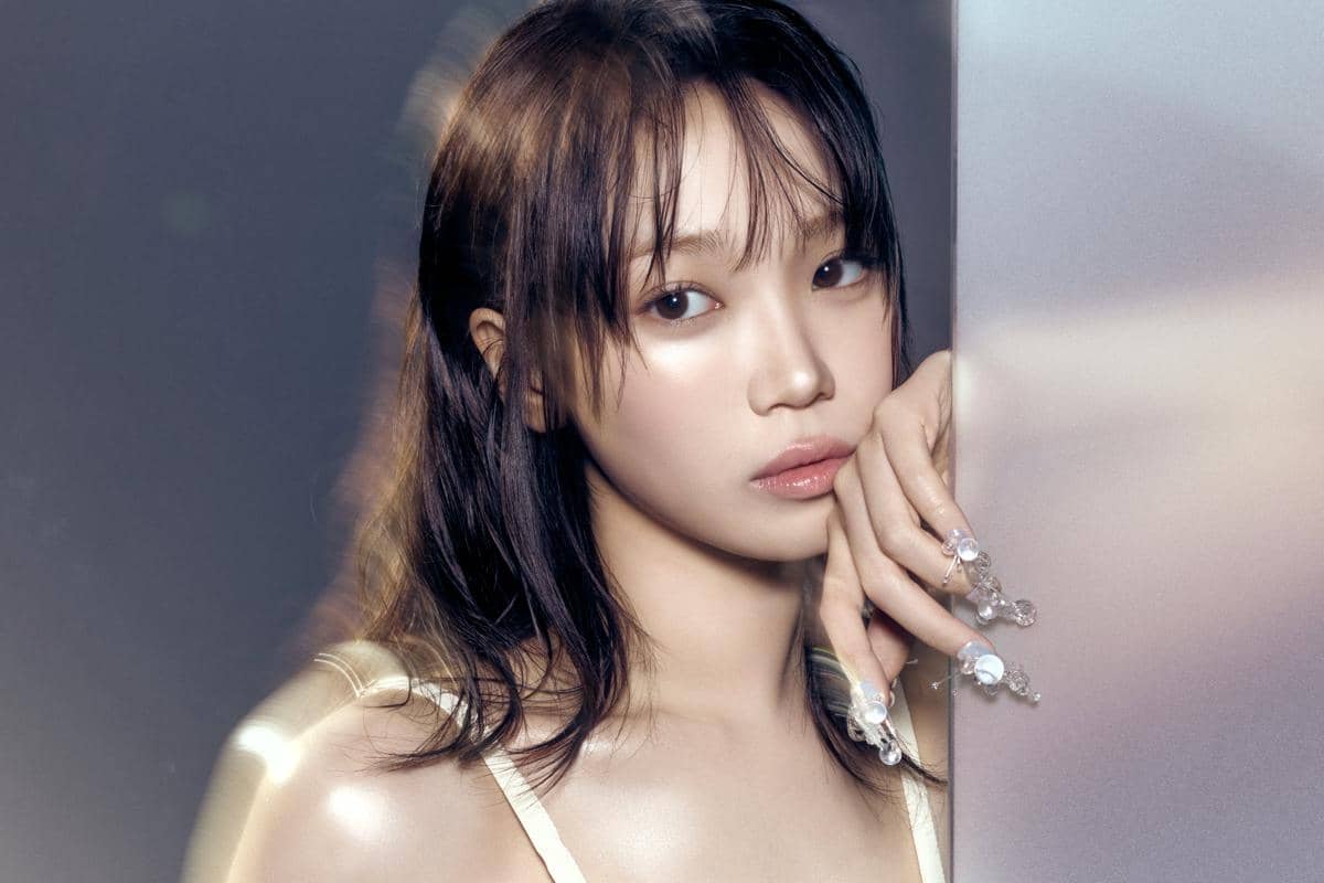 Seorang perempuan berpose dengan ekspresi lembut, rambut panjang berwarna gelap, dan kuku dihiasi ornamen kristal di latar lembut berwarna abu-abu.