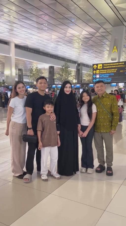 Sebuah keluarga berpose bersama di area keberangkatan bandara, dengan suasana ramai dan pencahayaan terang di sekitarnya.