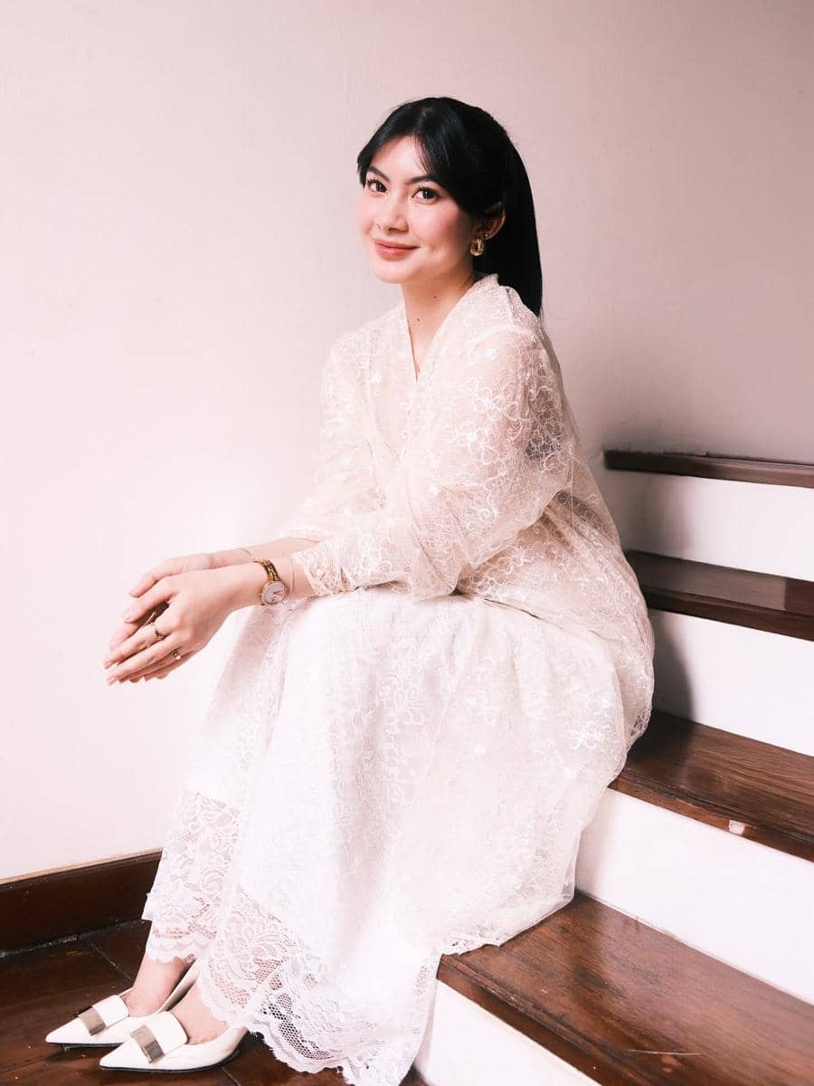 Seorang wanita berambut hitam mengenakan gaun renda putih duduk di tangga kayu sambil tersenyum ke arah kamera.