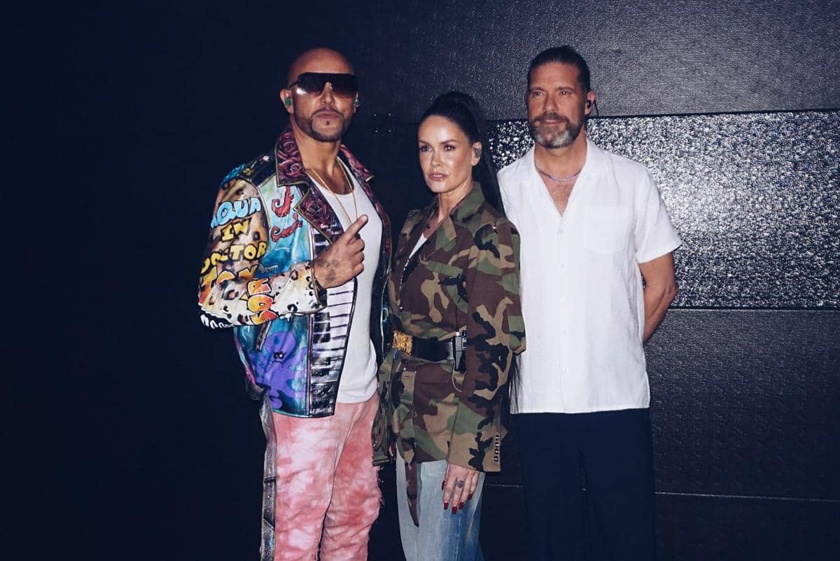 Tiga anggota band Aqua berpose bersama di depan latar gelap, mengenakan pakaian bergaya unik dengan jaket warna-warni, motif kamuflase, dan kemeja putih.