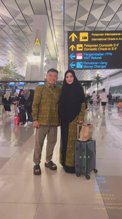 Sepasang suami istri mengenakan pakaian seragam batik berwarna hijau berfoto di bandara sebelum berangkat haji dengan koper di samping mereka.