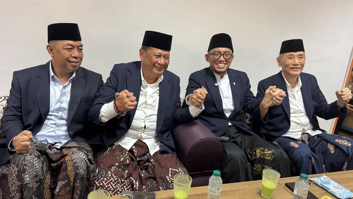 Delegasi Amirul Hajj Indonesia mengenakan jas dan sarung duduk berdampingan sambil bergandengan tangan dengan ekspresi gembira di ruang tunggu.