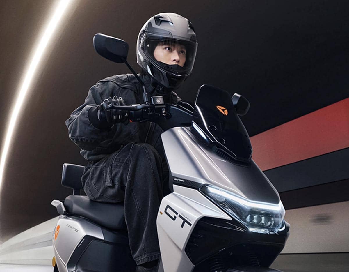 Seorang pengendara mengenakan helm dan jaket hitam sedang mengendarai skuter listrik Yadea GT berwarna abu-abu di terowongan modern.