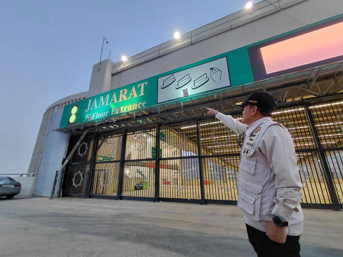 Petugas PPIH Arab Saudi mengenakan seragam krem menunjuk ke arah papan bertuliskan Jamarat 3rd Floor Entrance di area Mina.