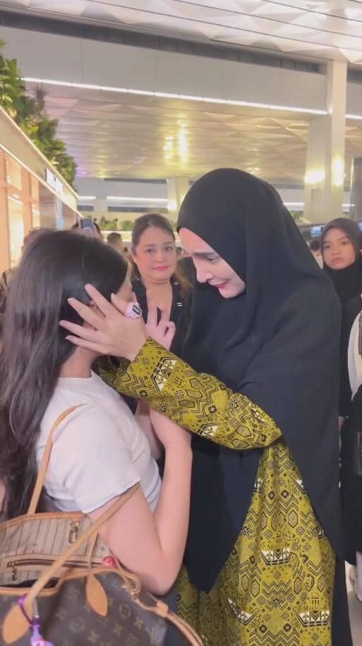 Seorang wanita berhijab hitam dan berpakaian kuning bermotif memeluk serta membelai wajah seorang gadis di bandara dengan suasana haru.