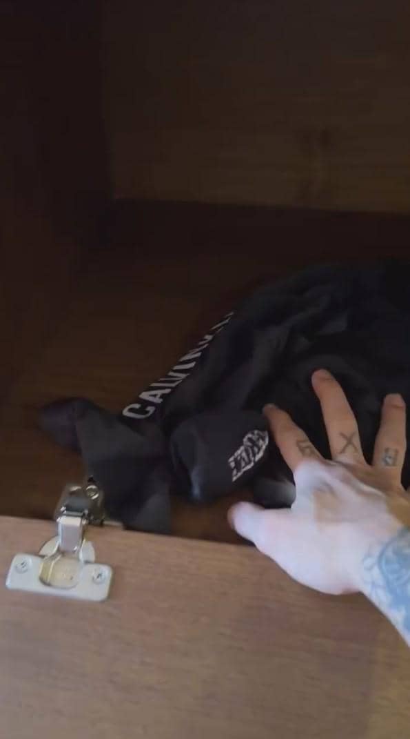 Tangan bertato sedang merapikan pakaian hitam bertulisan putih di dalam lemari kayu dengan engsel logam di bagian pintu.