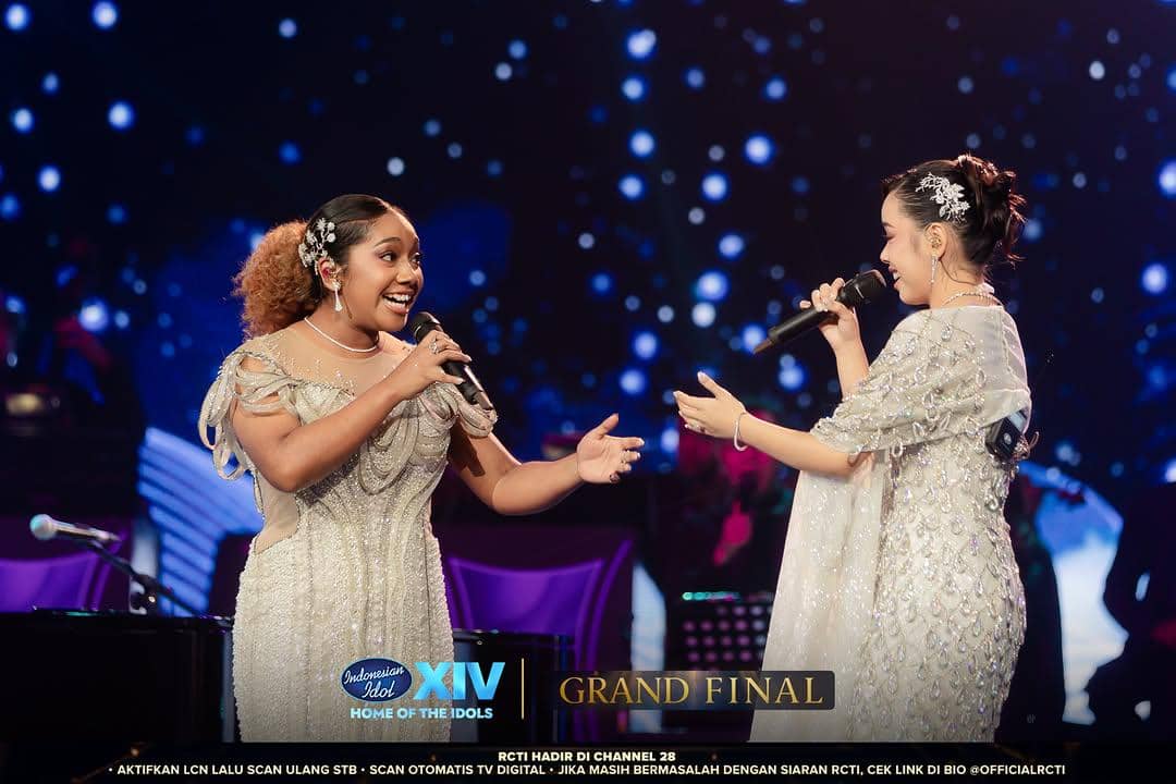 Dua finalis Indonesian Idol Season 14 bernyanyi bersama di panggung grand final dengan busana berkilau dan latar cahaya biru.