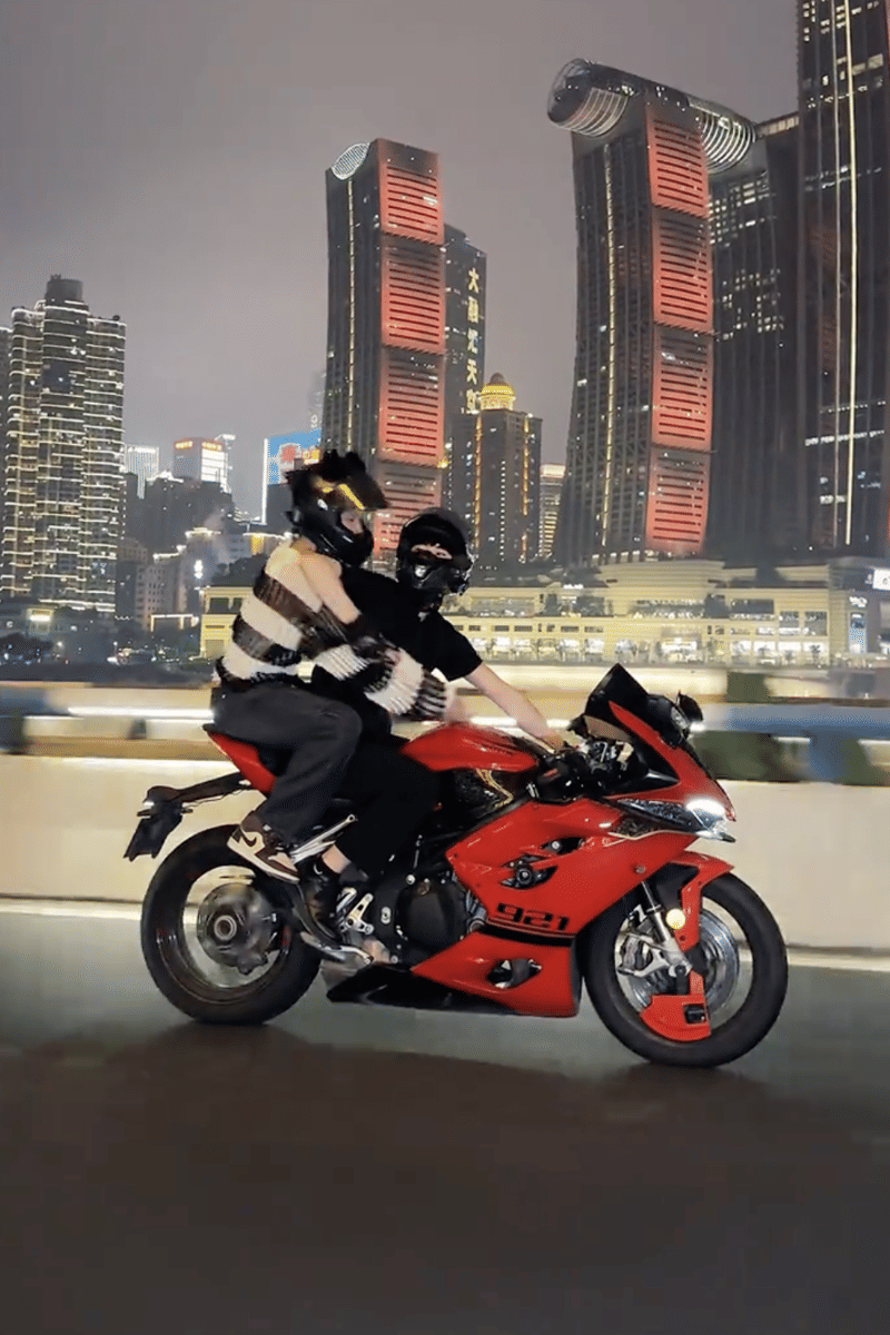 Dua orang mengenakan helm dan pakaian hitam berkendara dengan motor sport merah di jalan kota besar dengan gedung tinggi bercahaya malam hari.